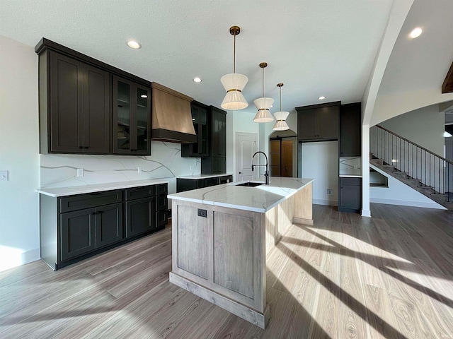 kitchen featuring custom range hood, decorative light fixtures, sink, an island with sink, and light hardwood / wood-style floors