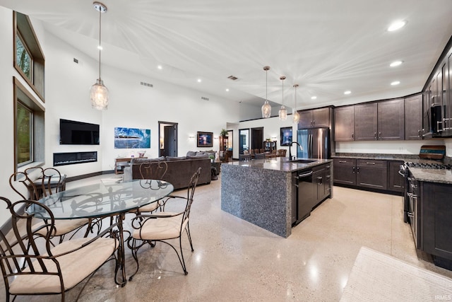 kitchen with stainless steel appliances, decorative light fixtures, dark stone counters, an island with sink, and a high ceiling