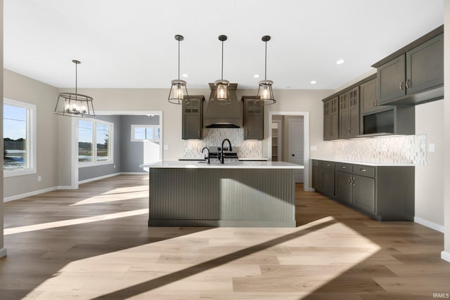 kitchen featuring premium range hood, dark hardwood / wood-style flooring, an island with sink, pendant lighting, and decorative backsplash