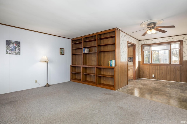 unfurnished living room with crown molding, carpet flooring, wooden walls, and ceiling fan