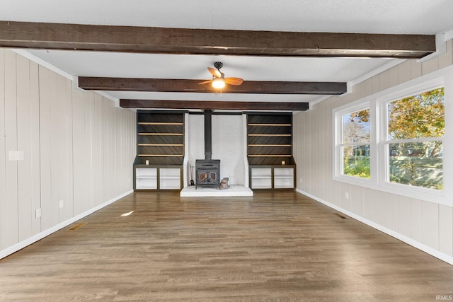 unfurnished living room with wooden walls, wood-type flooring, beamed ceiling, ceiling fan, and a wood stove