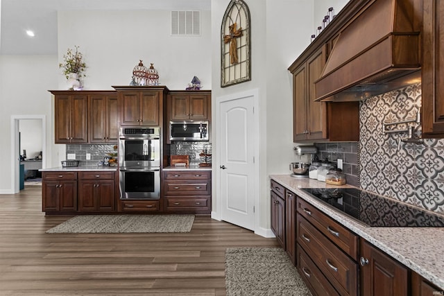 kitchen with light stone countertops, dark wood-type flooring, tasteful backsplash, premium range hood, and appliances with stainless steel finishes