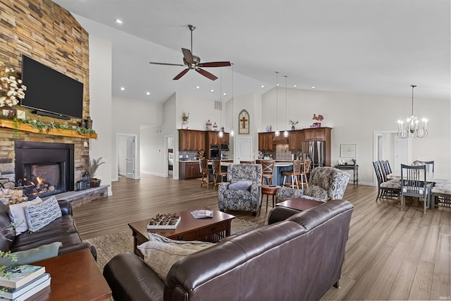 living room featuring a stone fireplace, high vaulted ceiling, light hardwood / wood-style floors, and ceiling fan with notable chandelier