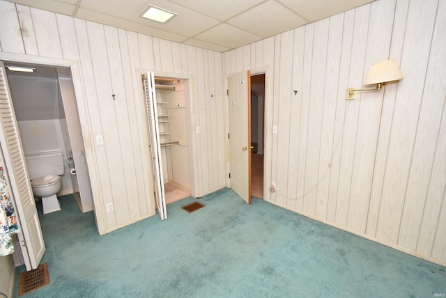 unfurnished bedroom featuring ensuite bathroom, a drop ceiling, carpet floors, a closet, and wood walls