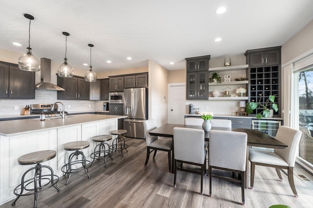 kitchen featuring an island with sink, stainless steel appliances, hanging light fixtures, and wall chimney range hood