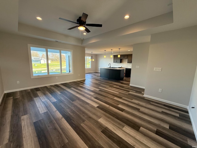 unfurnished living room with a tray ceiling, sink, dark wood-type flooring, and ceiling fan with notable chandelier