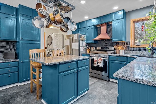 kitchen with stacked washer and dryer, blue cabinetry, stainless steel appliances, and wall chimney range hood