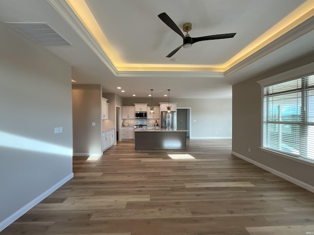 kitchen with hanging light fixtures, a raised ceiling, stainless steel appliances, a kitchen island with sink, and white cabinets