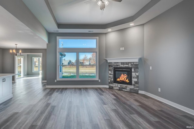 unfurnished living room with ceiling fan with notable chandelier, a stone fireplace, wood-type flooring, and a tray ceiling