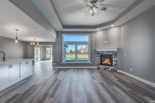 unfurnished living room featuring ceiling fan with notable chandelier, a raised ceiling, dark wood-type flooring, sink, and a stone fireplace