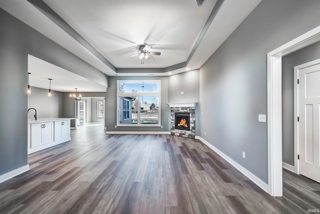 unfurnished living room featuring sink, dark wood-type flooring, a raised ceiling, a fireplace, and ceiling fan with notable chandelier