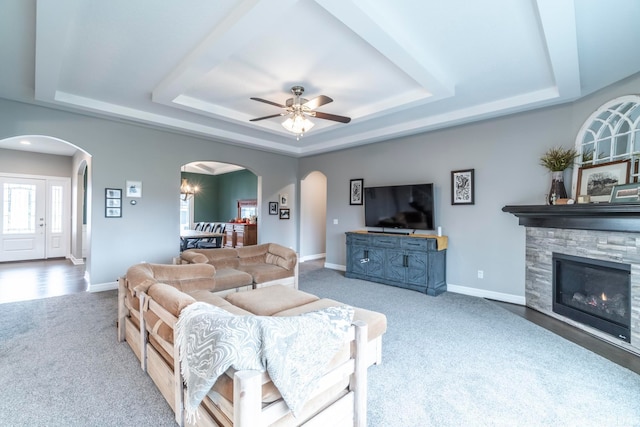 living room with a tray ceiling, a stone fireplace, carpet, and ceiling fan with notable chandelier