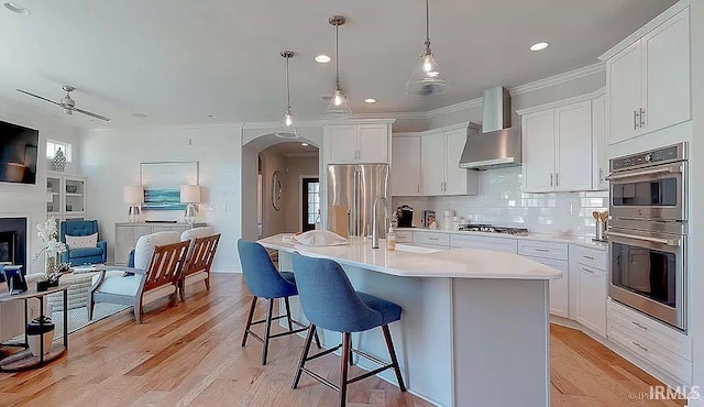 kitchen featuring ceiling fan, wall chimney exhaust hood, a center island with sink, white cabinets, and appliances with stainless steel finishes