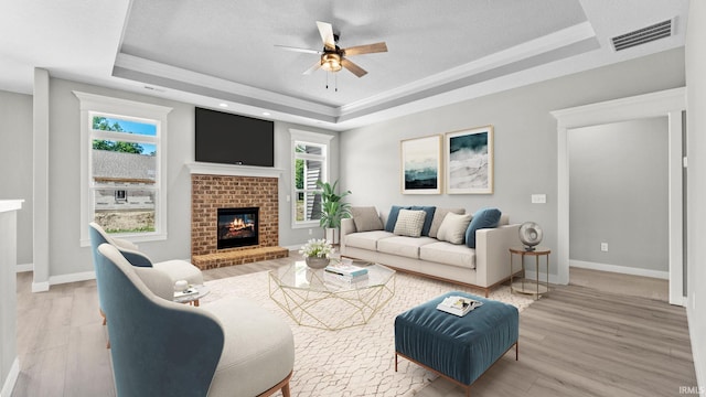 living room with ceiling fan, light hardwood / wood-style flooring, a tray ceiling, and a brick fireplace