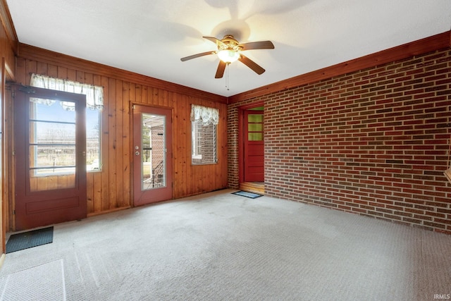 empty room with carpet, wood walls, ceiling fan, ornamental molding, and brick wall