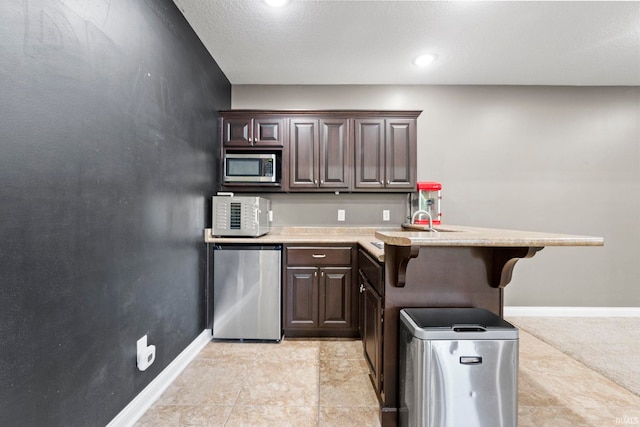 kitchen with kitchen peninsula, a textured ceiling, dark brown cabinetry, stainless steel appliances, and a breakfast bar area