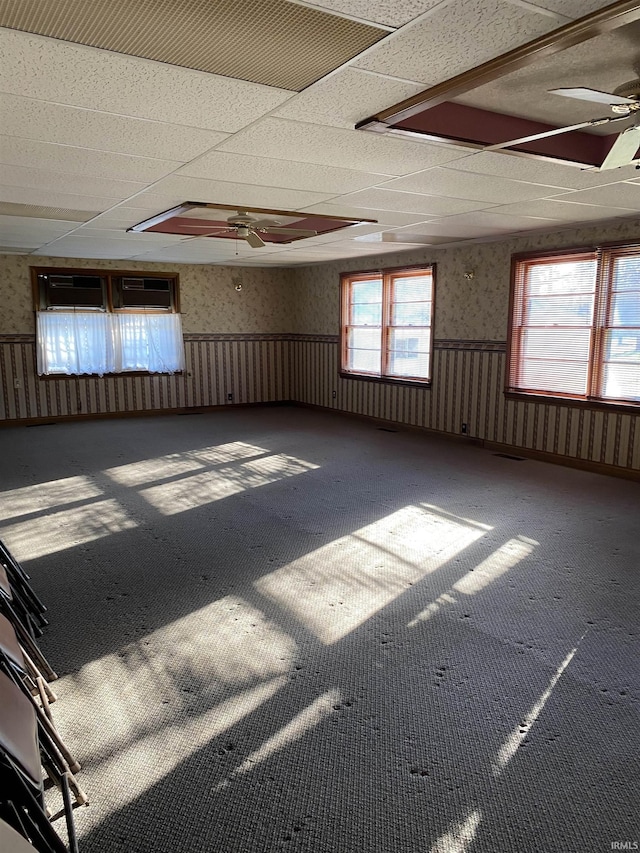 carpeted empty room featuring a wainscoted wall, a drop ceiling, a ceiling fan, and wallpapered walls