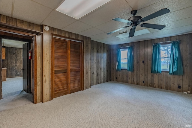 unfurnished bedroom with carpet flooring, ceiling fan, a paneled ceiling, and wooden walls