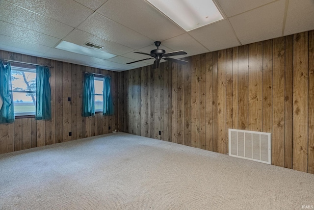 carpeted empty room with a paneled ceiling, wood walls, and ceiling fan