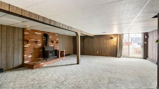 basement featuring a drop ceiling, wood walls, a wood stove, and carpet floors