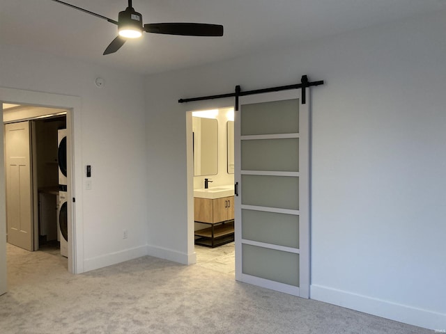 unfurnished bedroom featuring ensuite bath, ceiling fan, a barn door, stacked washing maching and dryer, and light colored carpet