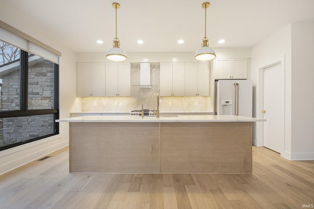 kitchen featuring white cabinets, white refrigerator with ice dispenser, wall chimney range hood, and light wood-type flooring