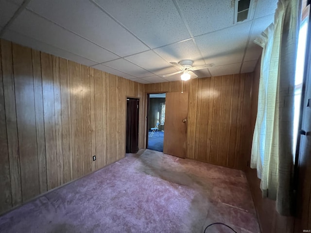 carpeted empty room featuring a paneled ceiling, wooden walls, and ceiling fan