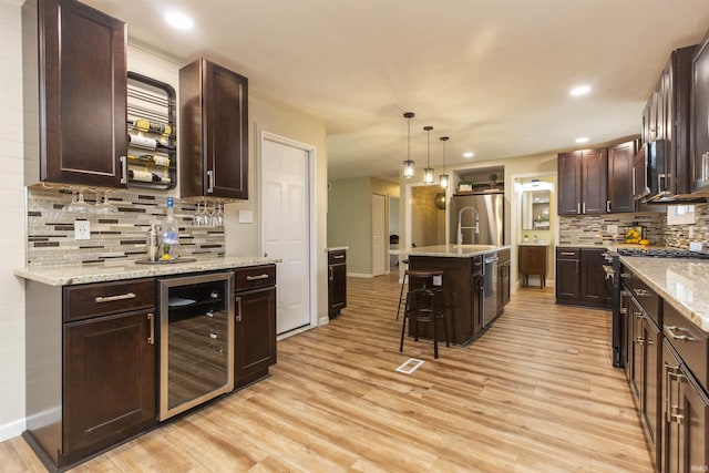 kitchen featuring beverage cooler, pendant lighting, light wood-type flooring, a kitchen island with sink, and appliances with stainless steel finishes