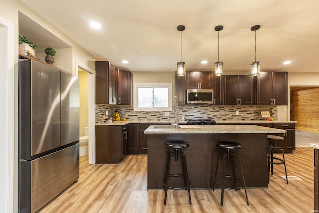 kitchen with appliances with stainless steel finishes, dark brown cabinets, pendant lighting, a center island with sink, and light hardwood / wood-style flooring