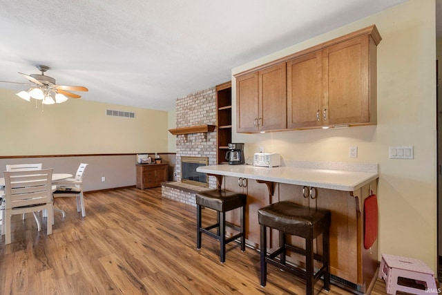 kitchen featuring ceiling fan, a brick fireplace, kitchen peninsula, a kitchen bar, and light wood-type flooring