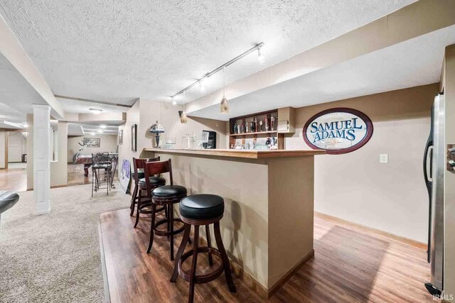 bar featuring rail lighting, stainless steel fridge, wood-type flooring, a textured ceiling, and decorative light fixtures