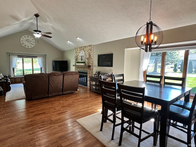 dining area with ceiling fan with notable chandelier, a stone fireplace, vaulted ceiling, a textured ceiling, and wood-type flooring