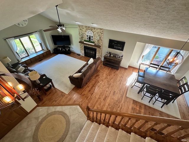 living room featuring ceiling fan with notable chandelier, a textured ceiling, vaulted ceiling, and a stone fireplace