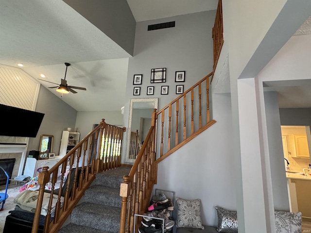 staircase with carpet, a tiled fireplace, ceiling fan, and high vaulted ceiling