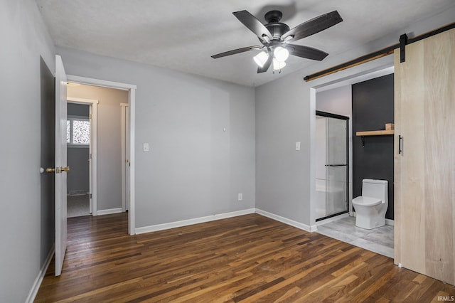 unfurnished bedroom featuring dark hardwood / wood-style flooring, a barn door, ensuite bathroom, and ceiling fan