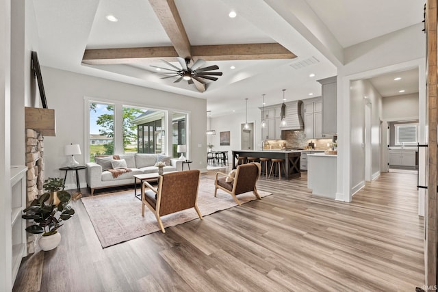 living room featuring a stone fireplace, ceiling fan, beamed ceiling, and light hardwood / wood-style floors