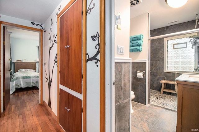 bathroom featuring vanity, tiled shower, wood-type flooring, tile walls, and toilet