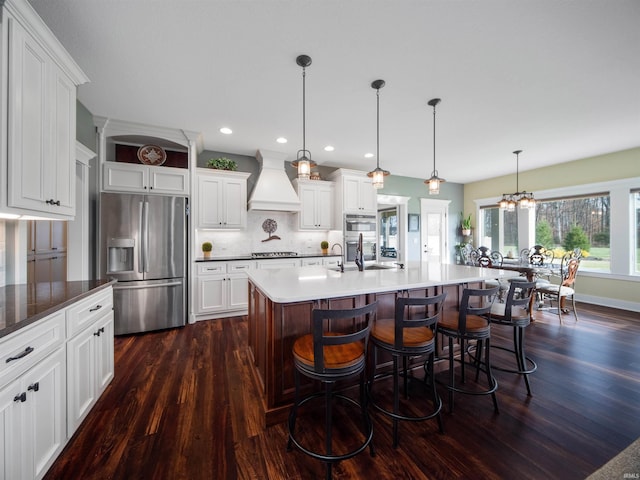 kitchen with appliances with stainless steel finishes, custom range hood, pendant lighting, a notable chandelier, and an island with sink