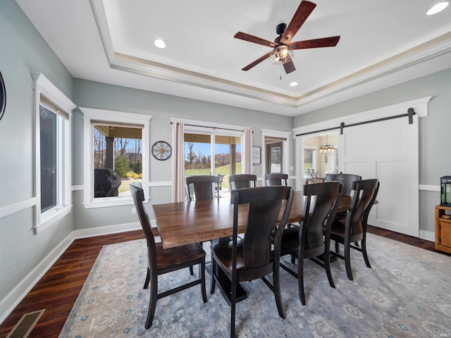 dining space with ceiling fan, dark wood-type flooring, a raised ceiling, a barn door, and ornamental molding