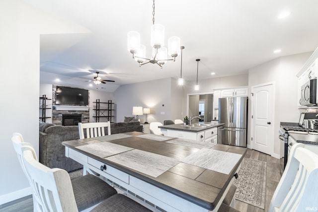 dining area featuring dark hardwood / wood-style flooring, ceiling fan with notable chandelier, vaulted ceiling, sink, and a stone fireplace