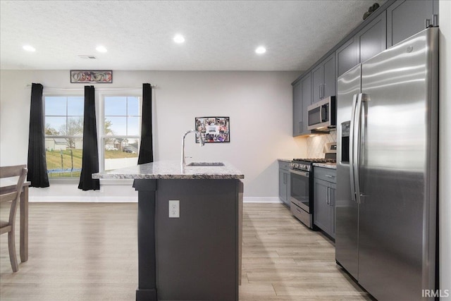 kitchen featuring appliances with stainless steel finishes, light stone counters, a textured ceiling, sink, and a center island with sink