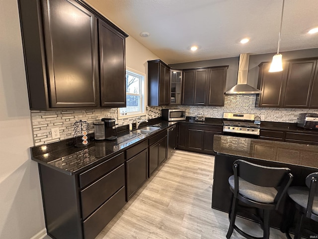 kitchen featuring appliances with stainless steel finishes, wall chimney exhaust hood, dark brown cabinetry, decorative light fixtures, and dark stone countertops