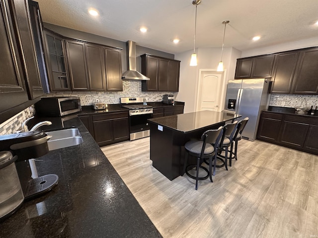 kitchen featuring wall chimney exhaust hood, decorative light fixtures, a kitchen island, a kitchen bar, and stainless steel appliances