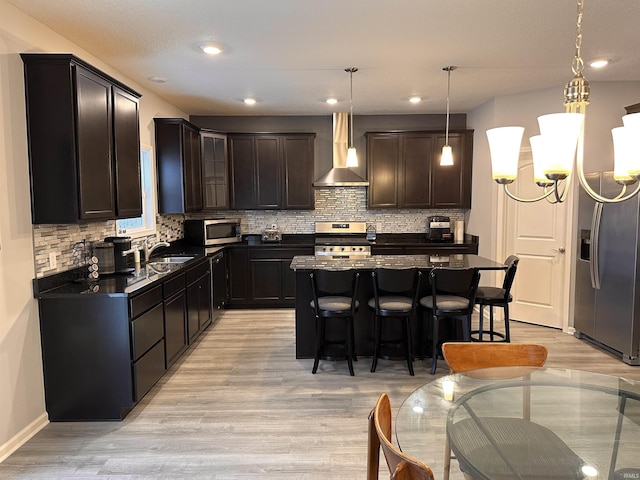 kitchen with appliances with stainless steel finishes, wall chimney exhaust hood, decorative light fixtures, light hardwood / wood-style floors, and a kitchen island