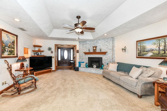 living room with ceiling fan, a brick fireplace, a raised ceiling, a textured ceiling, and carpet