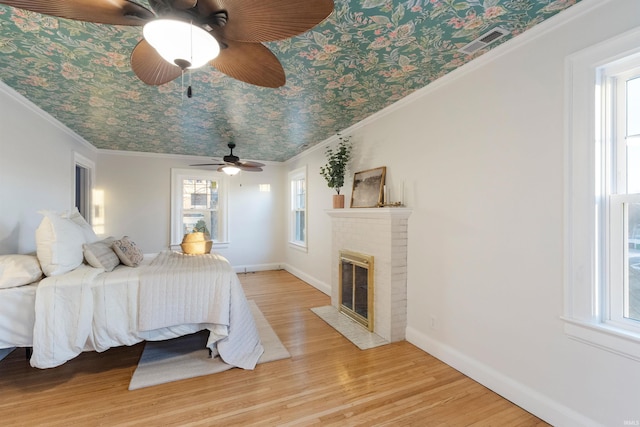 bedroom with ceiling fan, a brick fireplace, crown molding, multiple windows, and light wood-type flooring