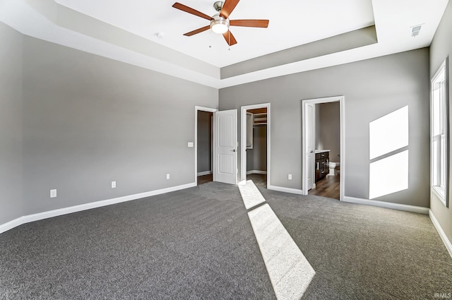 unfurnished bedroom with ensuite bath, a spacious closet, ceiling fan, dark colored carpet, and a raised ceiling