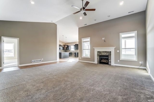 unfurnished living room featuring carpet flooring, ceiling fan, a stone fireplace, and a wealth of natural light