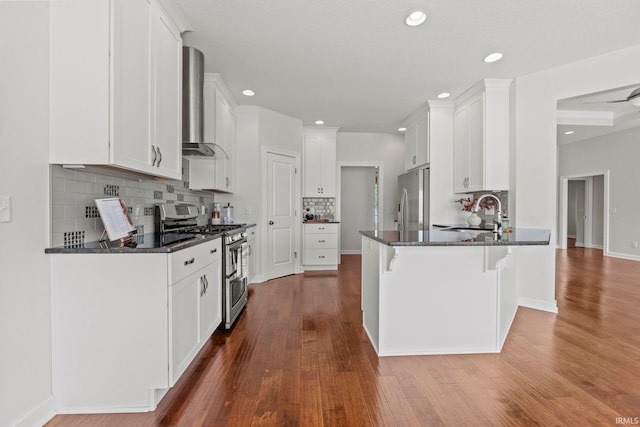 kitchen featuring kitchen peninsula, dark stone counters, wall chimney exhaust hood, stainless steel appliances, and white cabinetry