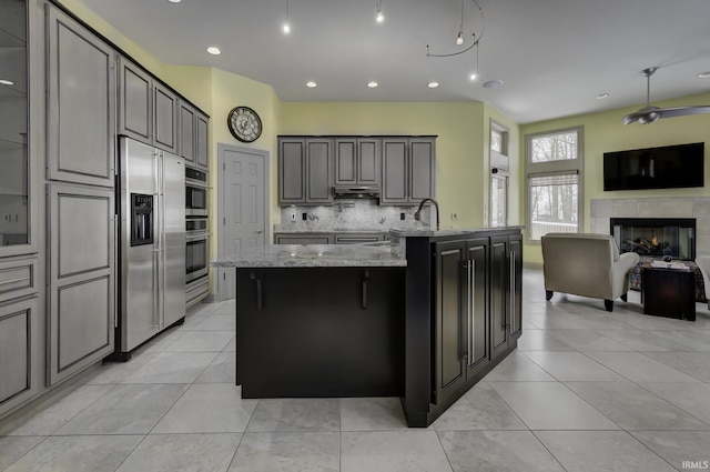 kitchen with a center island with sink, decorative backsplash, light tile patterned floors, light stone counters, and a tiled fireplace